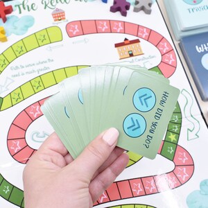 K&ouml;nnte beinhalten: Eine Hand h&auml;lt einen Stapel gr&uuml;ner Karten mit blauen Pfeilen auf der obersten Karte. Die Karte lautet "Wie hast du das gemacht?" Die Karten liegen auf einem Spielbrett mit einem roten und gr&uuml;nen Pfad und Sternen.