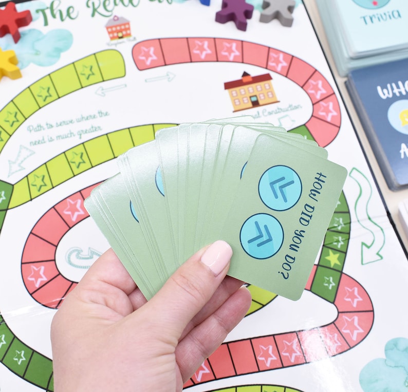 K&ouml;nnte beinhalten: Eine Hand h&auml;lt einen Stapel gr&uuml;ner Karten mit blauen Pfeilen auf der obersten Karte. Die Karte lautet "Wie hast du das gemacht?" Die Karten liegen auf einem Spielbrett mit einem roten und gr&uuml;nen Pfad und Sternen.