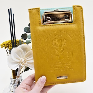 May include: A yellow leather clipboard with a gold metal clip and a gold embossed design. The design features a tree in a pot with the text "continued without letup declaring good news" and "best life ever".