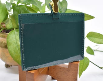 May include: A dark green leather badge holder with a gold clip. The holder has a clear plastic window for displaying a badge or ID card.