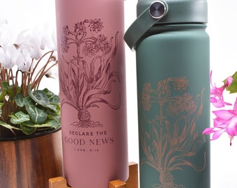 May include: Two stainless steel water bottles with a floral design and the text "Declare the Good News 1 Cor. 9:16". The bottles are pink and green and are on a wooden stand.