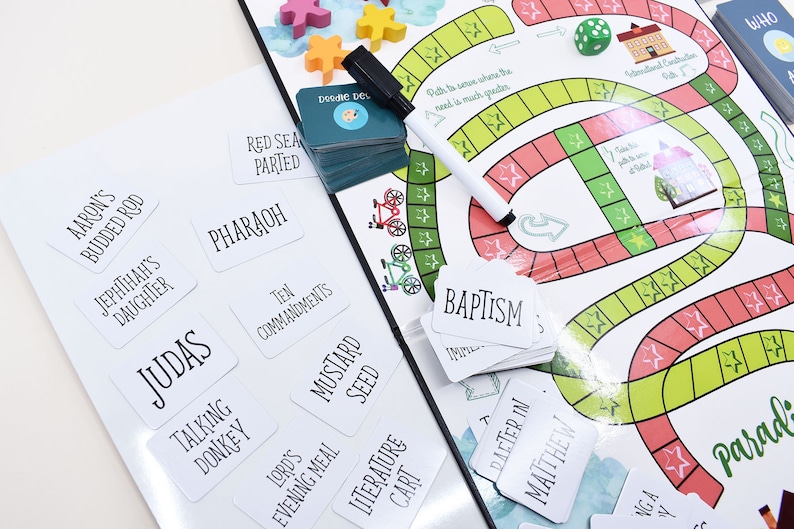 K&ouml;nnte beinhalten: Ein wei&szlig;es Brettspiel mit einer bunten Bahn und einem Stapel wei&szlig;er Karten mit schwarzem Text. Die Karten haben W&ouml;rter wie "Pharao", "Zehn Gebote", "Judas" und "Taufe".
