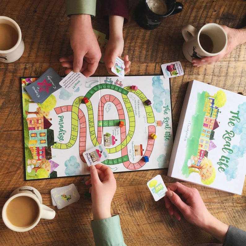 K&ouml;nnte beinhalten: Eine Familie spielt ein Brettspiel namens "The Real Life" mit bunten H&auml;usern, einem gewundenen Pfad und Karten mit S&auml;tzen wie "Paradies" und "Schulden".