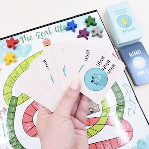 K&ouml;nnte beinhalten: Ein Brettspiel namens "The Real Life" mit einem farbenfrohen Spielbrett, Spielfiguren und Karten. Die Karten haben ein Musiksymbol und das Wort "Hum" darauf geschrieben.