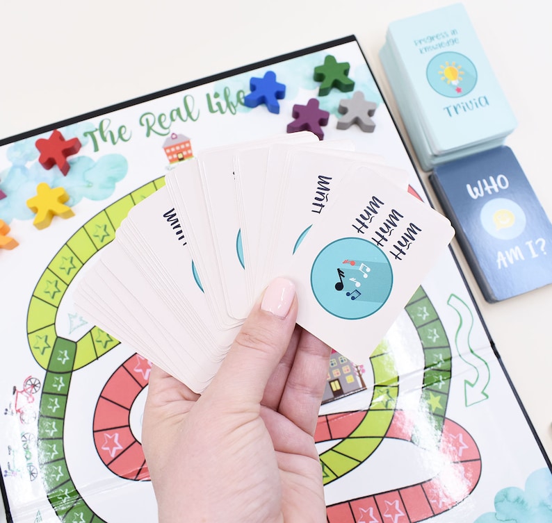 K&ouml;nnte beinhalten: Ein Brettspiel namens "The Real Life" mit einem farbenfrohen Spielbrett, Spielfiguren und Karten. Die Karten haben ein Musiksymbol und das Wort "Hum" darauf geschrieben.
