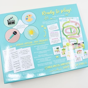 K&ouml;nnte beinhalten: Eine blaue und gr&uuml;ne Spielschachtel mit dem Titel "Ready to play!" und dem Text "Land on a star". Die Schachtel zeigt Illustrationen einer Filmklappe, einer Palette, eines Mikrofons, einer Gl&uuml;hbirne und eines Spielbretts mit einem Pfad und H&auml;usern. Der Text "Deluxe version: New Includes" ist ebenfalls sichtbar.