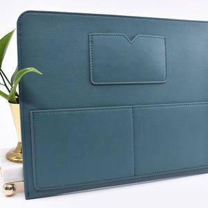 May include: Teal-colored document holder with a textured finish. Features a small pocket with a V-shaped top and two larger pockets below. The holder is rectangular and appears to be made of leather or a similar material.