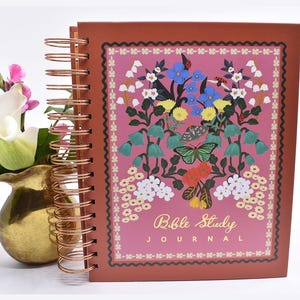 May include: A spiral-bound Bible Study Journal with a copper-colored wire coil. The cover is a deep terracotta shade featuring a central pink rectangle decorated with colorful illustrations of flowers, butterflies, and ladybugs, with the text Bible Study JOURNAL printed in gold.
