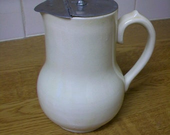 May include: A cream-colored ceramic pitcher with a rounded body and a curved handle. The pitcher features a hinged, circular metal lid with a small white knob at the center, designed to cover the opening and the pouring spout securely.