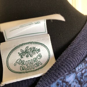 May include: A white clothing label with a green oval border and the text Laura Ashley in green, featuring a floral graphic above the text. A smaller white tag above it reads 100% cotton/coton/katoen/baumwolle. The garment has a dark blue ribbed collar.