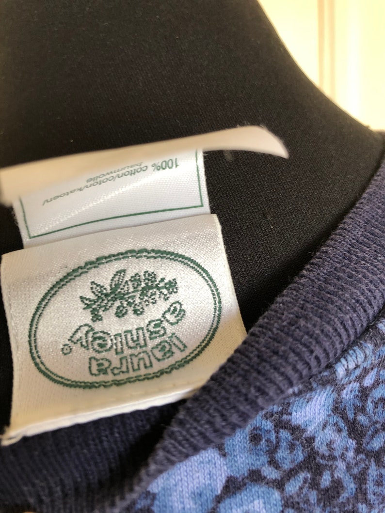 May include: A white clothing label with a green oval border and the text Laura Ashley in green, featuring a floral graphic above the text. A smaller white tag above it reads 100% cotton/coton/katoen/baumwolle. The garment has a dark blue ribbed collar.