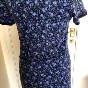 May include: A short-sleeved dress featuring a dense, all-over floral pattern in shades of light blue and periwinkle against a dark navy blue background. The garment includes a horizontal seam across the waist and dark navy blue trim on the sleeves.
