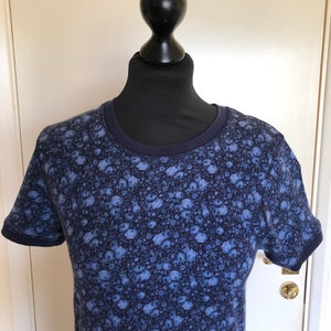May include: A short-sleeved t-shirt featuring a dark blue and light blue floral pattern throughout. The garment has a solid dark blue ribbed crew neckline and matching dark blue ribbed sleeve cuffs, providing a contrast to the overall floral print.