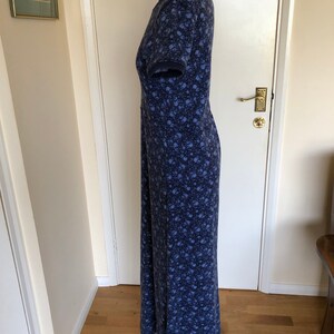 May include: A floor-length dress with short sleeves, featuring a dark blue base color and a dense, repeating pattern of small, light blue floral motifs. The garment is displayed on a mannequin, showing a straight silhouette and a round neckline.