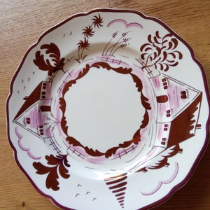 Könnte beinhalten: Ein weißer Keramikteller mit einem braunen und rosafarbenen Design. Das Design zeigt zwei Häuser mit Bäumen und einem Zaun. Der Teller hat einen gewellten Rand.