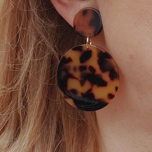 Può includere: Un paio di orecchini rotondi in tartaruga marrone e nera. Gli orecchini sono realizzati in un materiale liscio e lucido e hanno un perno dorato.