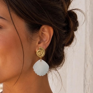 Peut inclure: Une boucle d'oreille dorée composée d'un clou texturé en forme de spirale sur le dessus et d'un pendentif en forme de coquillage blanc lisse en éventail suspendu en dessous. Le bijou présente une tige métallique et un élément pendant.