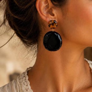 Può includere: Un orecchino pendente caratterizzato da un piccolo perno circolare con motivo tartarugato marrone e nero, collegato a un ciondolo circolare più grande, liscio e di colore nero lucido. L'orecchino è dotato di un semplice perno in metallo.