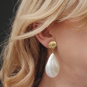 Könnte beinhalten: Goldfarbene Ohrringe mit einem muschelförmigen Stecker und einem großen, birnenförmigen, weißen Tropfen. Die Ohrringe werden am Ohr getragen, mit blondem Haar im Hintergrund. Ein klassisches Schmuckstück.