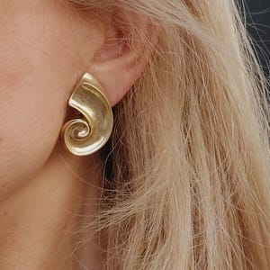 Könnte beinhalten: Goldfarbene Ohrringe in Spiralform. Die Ohrringe sind aus glattem, glänzendem Metall gefertigt und haben ein schlichtes, elegantes Design.