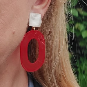 Può includere: Un paio di orecchini con un pendente ovale rosso e un pendente quadrato bianco. Gli orecchini sono in acrilico e hanno una finitura dorata.