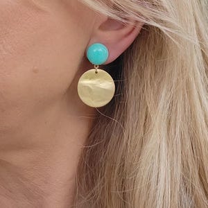 Pode incluir: Um par de brincos dourados com discos e uma esfera azul turquesa acima de cada disco. Os brincos estão pendurados na orelha de uma pessoa. Os brincos têm um design simples e elegante.