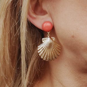Peut inclure: Boucles d'oreilles en forme de coquillage dorées avec une pierre pendante de couleur corail. Les boucles d'oreilles sont une pièce forte et seraient parfaites pour une fête d'été ou une journée à la plage.