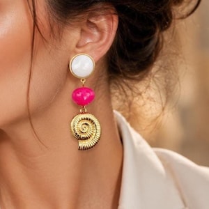 Könnte beinhalten: Ein Ohrhänger bestehend aus einem runden weißen Ohrstecker, einer glatten, pinkfarbenen Perle und einem goldfarbenen Metallanhänger in Form einer spiralförmigen Muschel. Die Elemente sind durch kleine goldene Ringe verbunden.