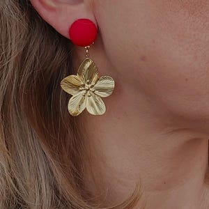May include: A close-up of a gold-colored flower-shaped earring with a red circular stud. The earring is attached to the ear. The flower has detailed petals and a textured surface, creating a striking contrast with the smooth red stud.