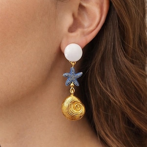 Peut inclure: Boucles d'oreilles pendantes avec un clou circulaire blanc, une étoile de mer bleue avec des accents bleus et un coquillage doré. Les boucles d'oreilles ont un thème plage ou océan.