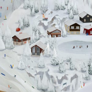 May include: An aerial view of a snowy ski resort scene. Miniature skiers descend slopes marked with colorful poles. Several snow-covered cabins and a frozen pond are nestled among snow-laden evergreen trees. A red cherry-shaped object is visible.