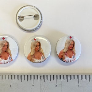 May include: Three white round button pins with a silver border. Each pin features a photo of a woman wearing a white nurse's uniform with a red cross on her hat. The woman is looking down at the camera. The pins are 1 inch in diameter.
