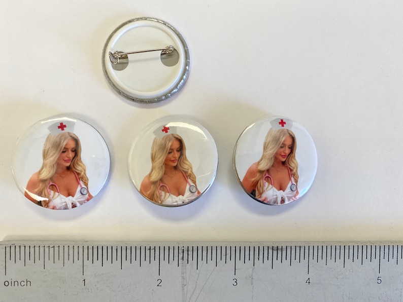 May include: Three white round button pins with a silver border. Each pin features a photo of a woman wearing a white nurse's uniform with a red cross on her hat. The woman is looking down at the camera. The pins are 1 inch in diameter.