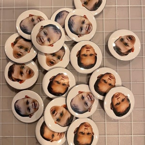 May include: A collection of 16 round white buttons with black borders, each featuring a different baby's face.