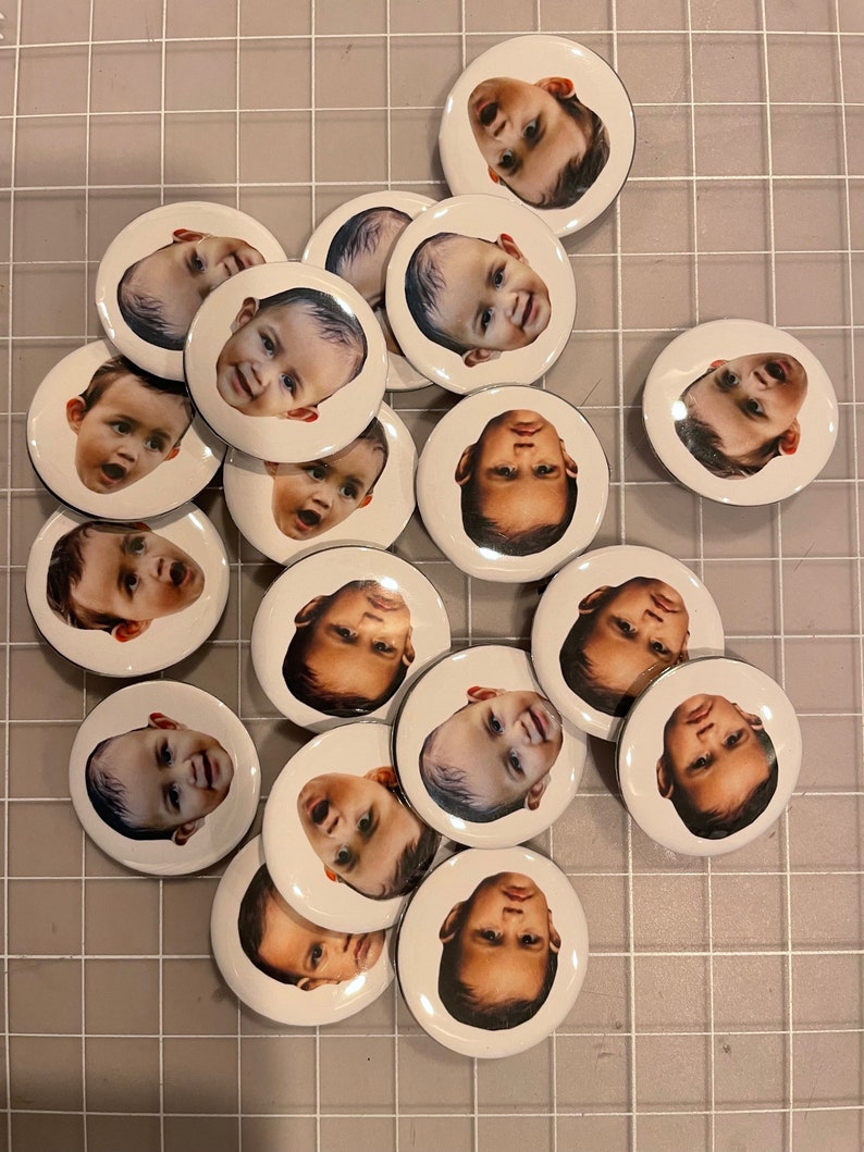 May include: A collection of 16 round white buttons with black borders, each featuring a different baby's face.