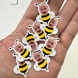 May include: A collection of nine bee-themed party favors with a child's face in the center of each bee. The bees are yellow and black with white wings and a white circle on each side of the bee.