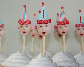 Puede incluir: Un conjunto de cupcakes decorados con sombreros de fiesta rojo, blanco y azul con la cara de un bebé en cada uno. Los sombreros tienen el número "1" en ellos.
