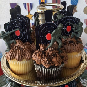 May include: A tiered silver serving stand with cupcakes decorated with chocolate frosting and military themed toppers. The cupcakes have black and white target paper with red bullseyes and green plastic army men figurines.