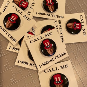 May include: A stack of white cards with black text that reads "Call Me" and a phone number "1-800-SUCCESS". Each card features a round button with a black background and a picture of a person wearing a red, white, and blue hat.