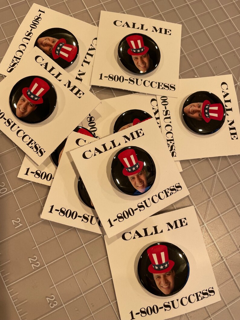 May include: A stack of white cards with black text that reads "Call Me" and a phone number "1-800-SUCCESS". Each card features a round button with a black background and a picture of a person wearing a red, white, and blue hat.