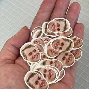 May include: A pile of round stickers with a person's face printed on each one. The person is smiling and has short brown hair.