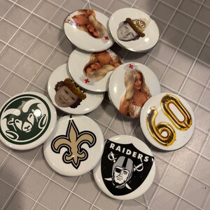 May include: A collection of round, white, and black buttons with various designs. Some buttons feature images of people, while others feature logos for sports teams, such as the New Orleans Saints and the Las Vegas Raiders. One button features a gold balloon with the number 60 on it.