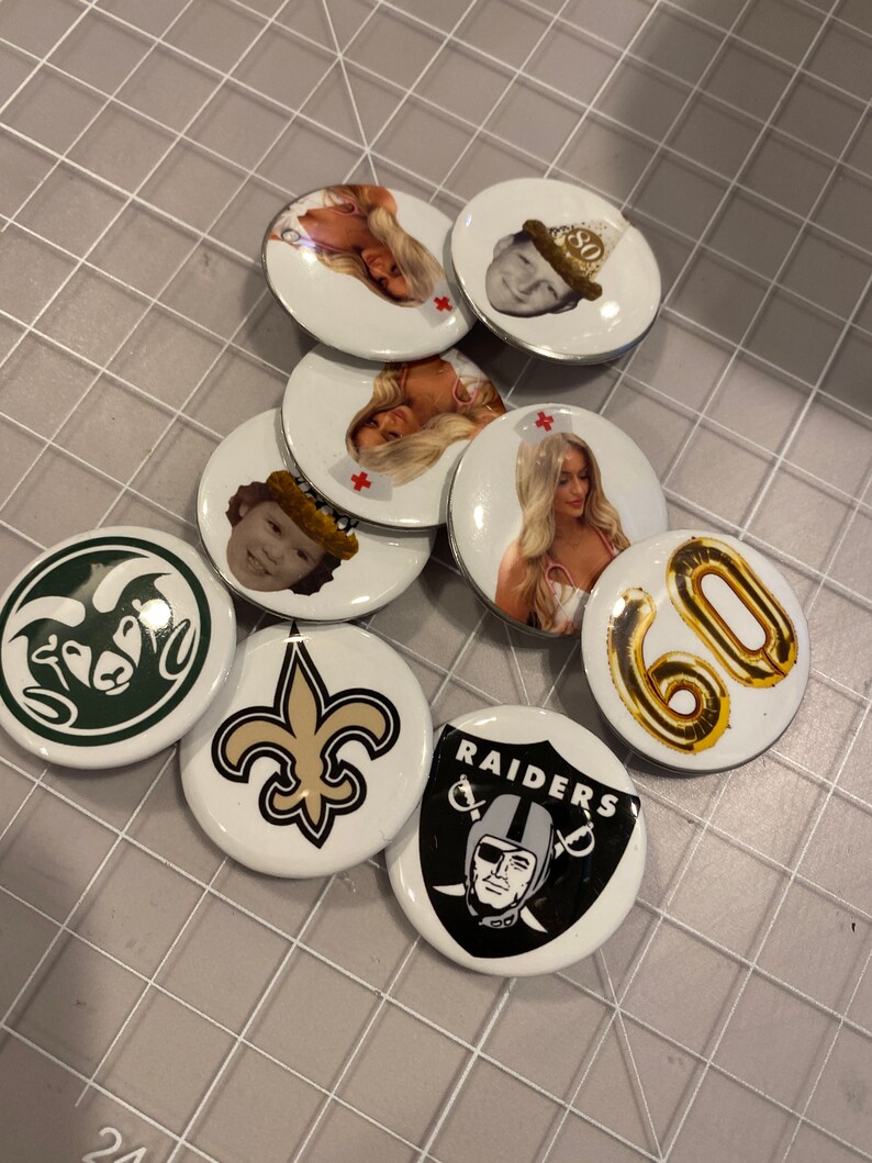 May include: A collection of round, white, and black buttons with various designs. Some buttons feature images of people, while others feature logos for sports teams, such as the New Orleans Saints and the Las Vegas Raiders. One button features a gold balloon with the number 60 on it.