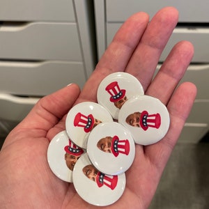 May include: A hand holding a pile of round, white buttons with a red, white, and blue design. Each button features a cartoon face wearing a red, white, and blue hat.