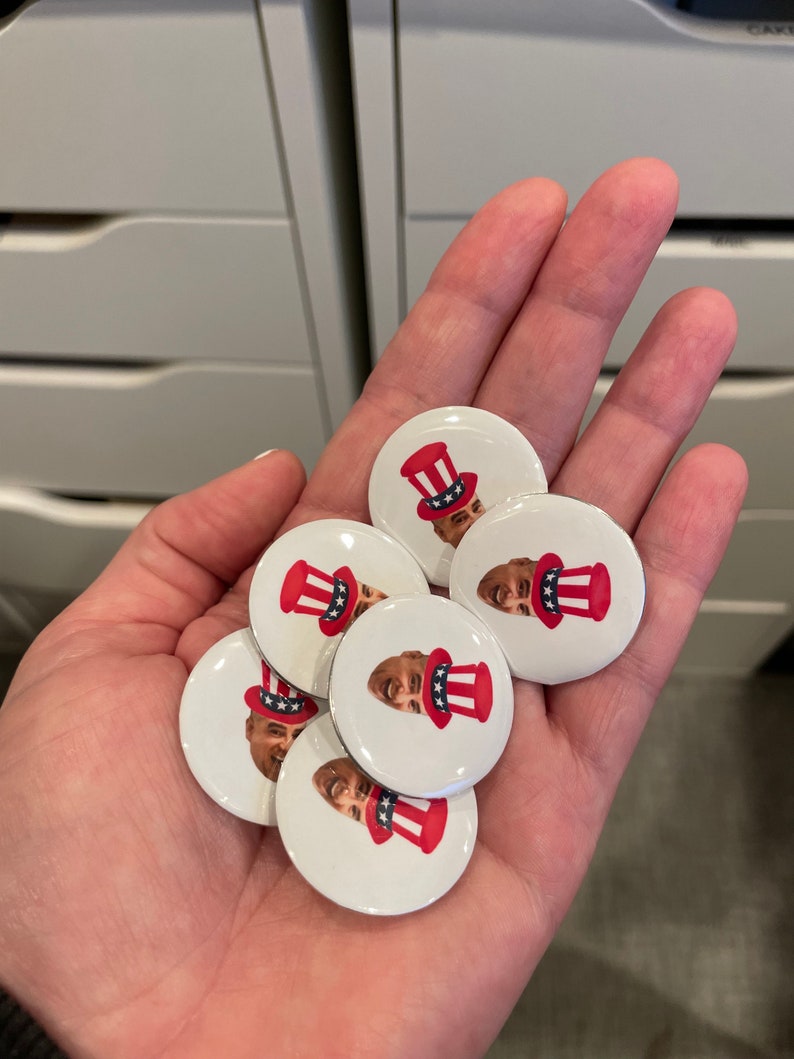 May include: A hand holding a pile of round, white buttons with a red, white, and blue design. Each button features a cartoon face wearing a red, white, and blue hat.