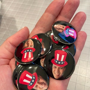 May include: A hand holding a pile of round, black and white buttons with a person's face and a red, white, and blue Uncle Sam hat on each button.