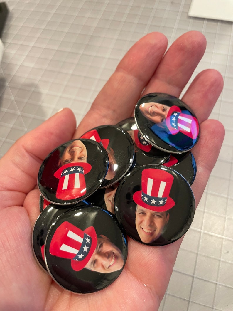 May include: A hand holding a pile of round, black and white buttons with a person's face and a red, white, and blue Uncle Sam hat on each button.