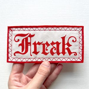 Könnte beinhalten: Ein rechteckiger Aufnäher mit dem Wort "Freak" in roter gotischer Schrift. Der Aufnäher hat einen weißen Hintergrund mit rotem Rand und roten Nähten. Der Aufnäher wird von einer Person in der Hand gehalten.