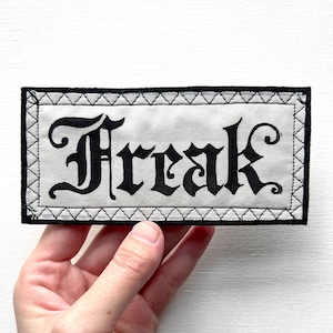 May include: A rectangular patch with the word "Freak" in black gothic lettering on a light gray background. The patch has a black border with white stitching and is held by a hand.