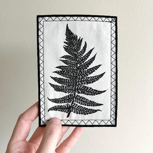 May include: Black and white embroidered fern leaf design on a white fabric patch with a black stitched border.